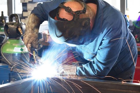 Man wearing welding helmet welding a piece of metal with cylinder of gas in the background